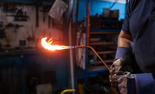 A welder wearing blue and grey work gloves holds out a flaming torch.